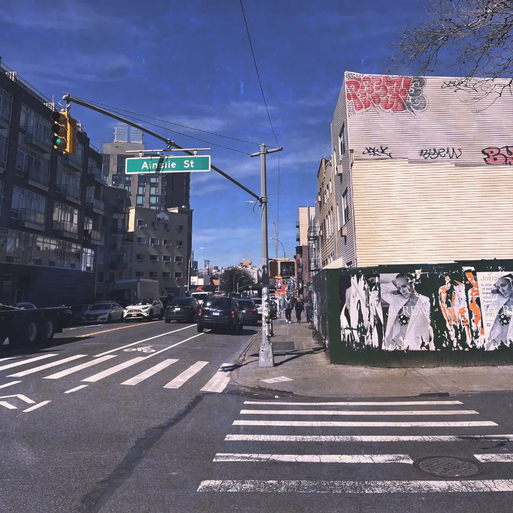 A vintage-styled photograph of a New York City street sign at an intersection; the sign reads 'Ainslie St'.