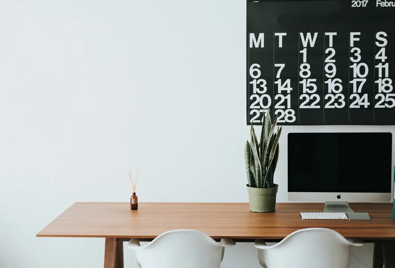 A photograph of a neatly organized desk workstation features the words 'Do More' on the monitor. In the background, a green houseplant hangs over a bookshelf.